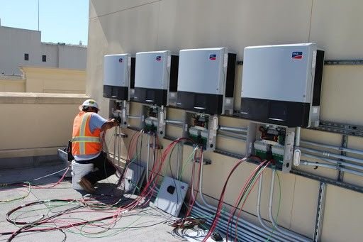 Solar panel inverter installation on a rooftop, with a worker in safety gear connecting wires to equipment.